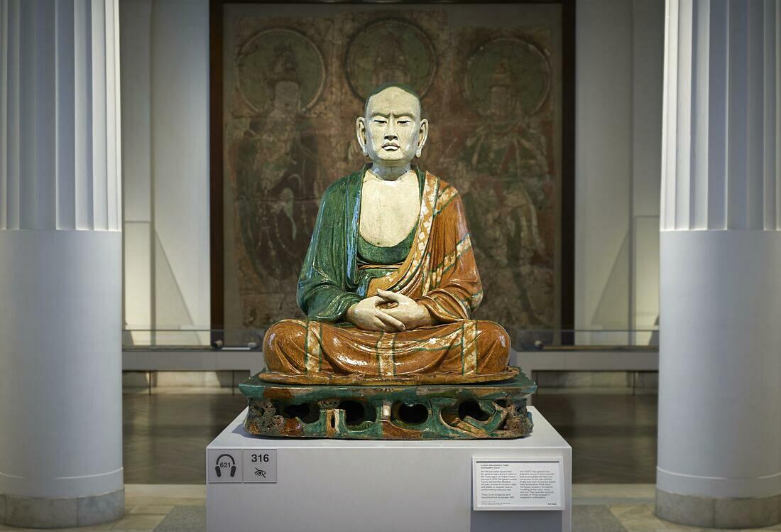 A statue of a meditating monk sitting on a pedestal. He wears yellow and green robes.