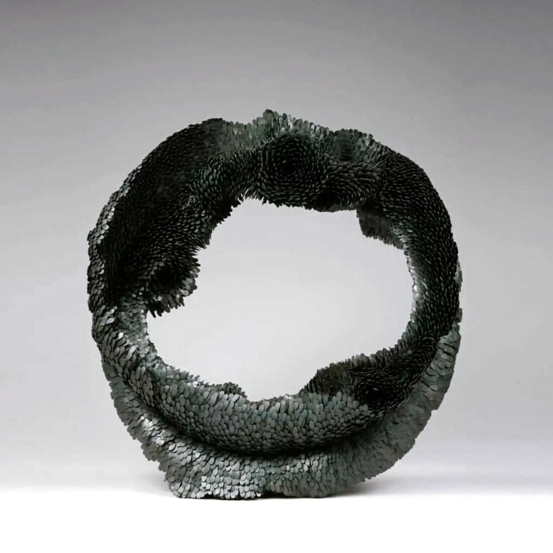 A black metal sculpture made of large amount of tiny petals creating a circle. The circle's surface is irregular as petals create flowering patterns at some places.