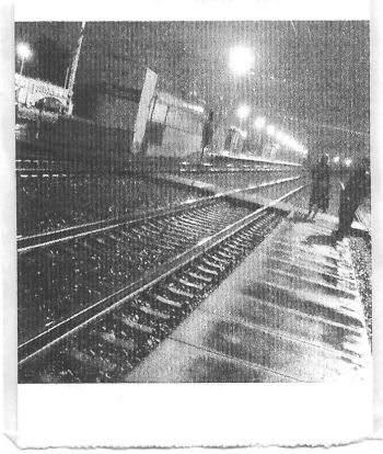 A black and white pocket printer style photo of a wet train platform before sunrise. There are light reflections on the trails and two people waiting.