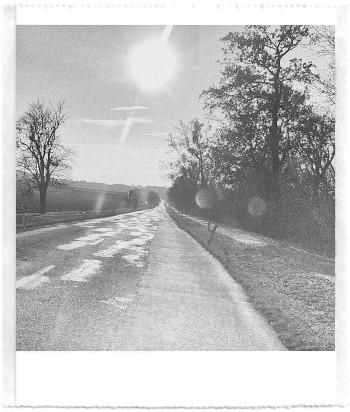 A black and white pocket printer style photo of sunlight reflections on patches of the road that is stretching far away.